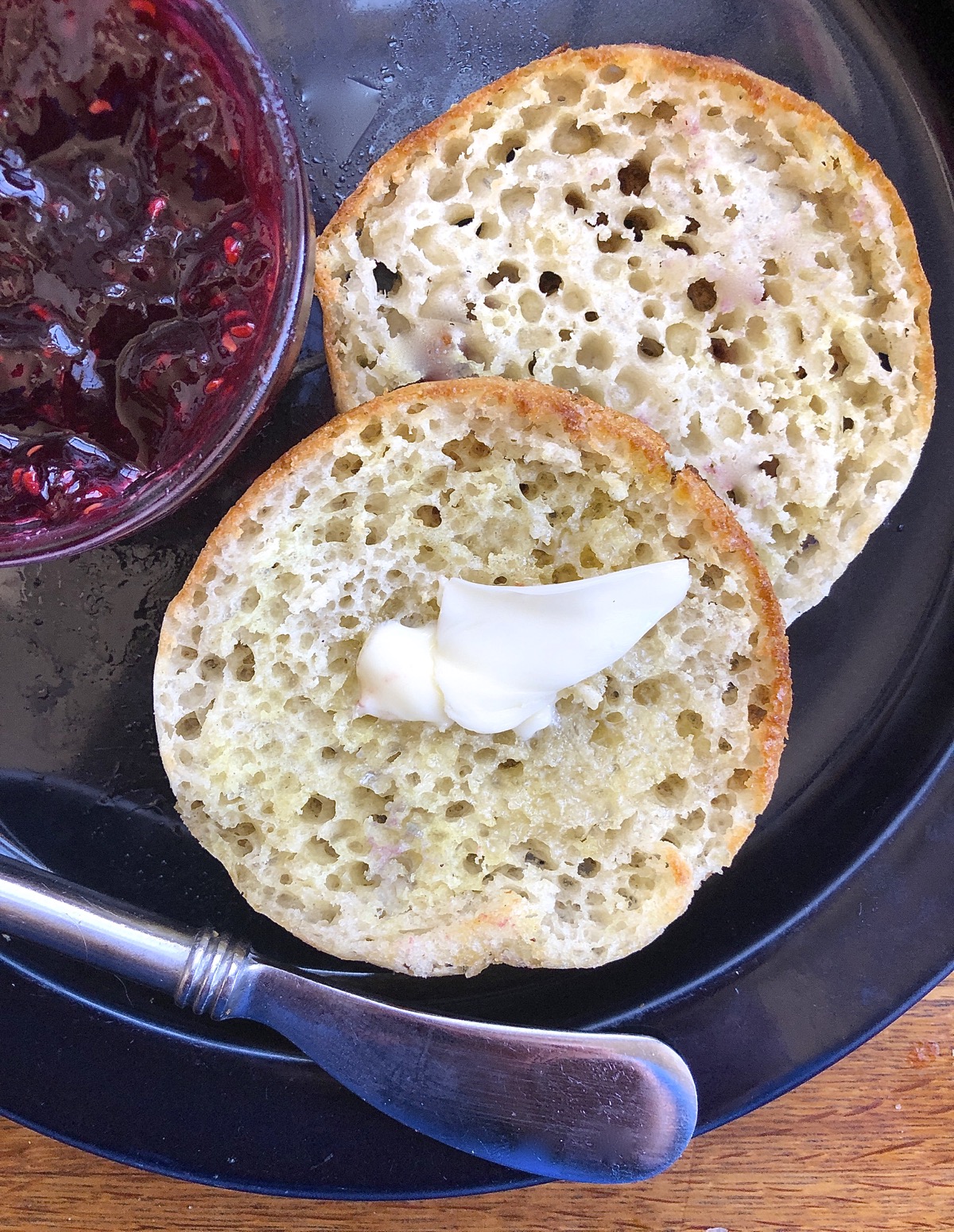 A sourdough crumpet split, warmed, and spread with butter, raspberry jam on the side.