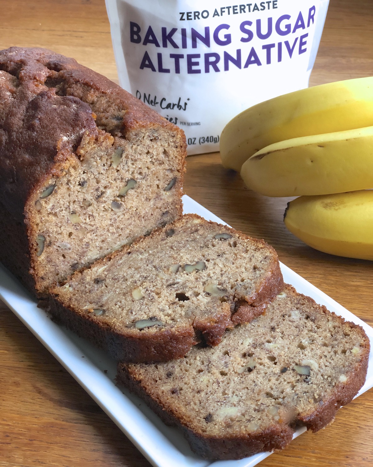 Loaf of banana bread on a plate, sliced, bananas and bag of Baking Sugar Alternative in the background.