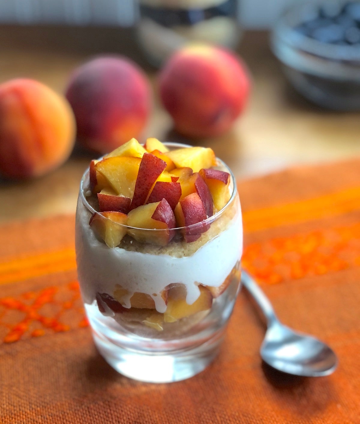 A peach dessert in a clear cocktail glass showing layers of cookies, diced peaches and whipped cream.