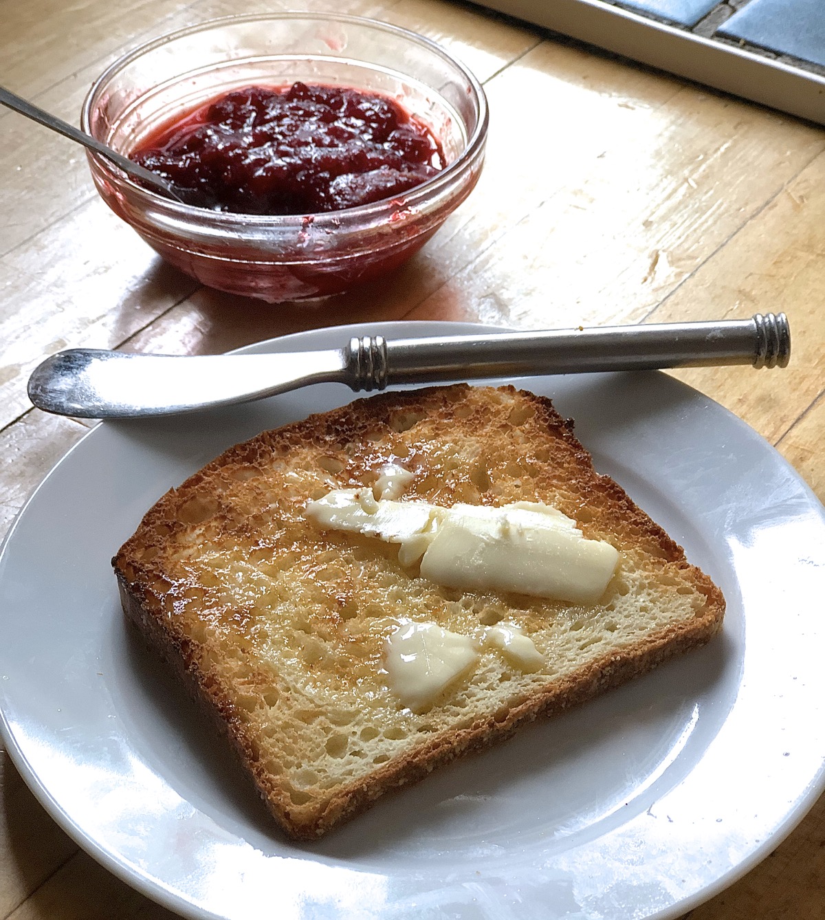 Piece of buttered toast on a plate, jar of strawberry preserves in background.