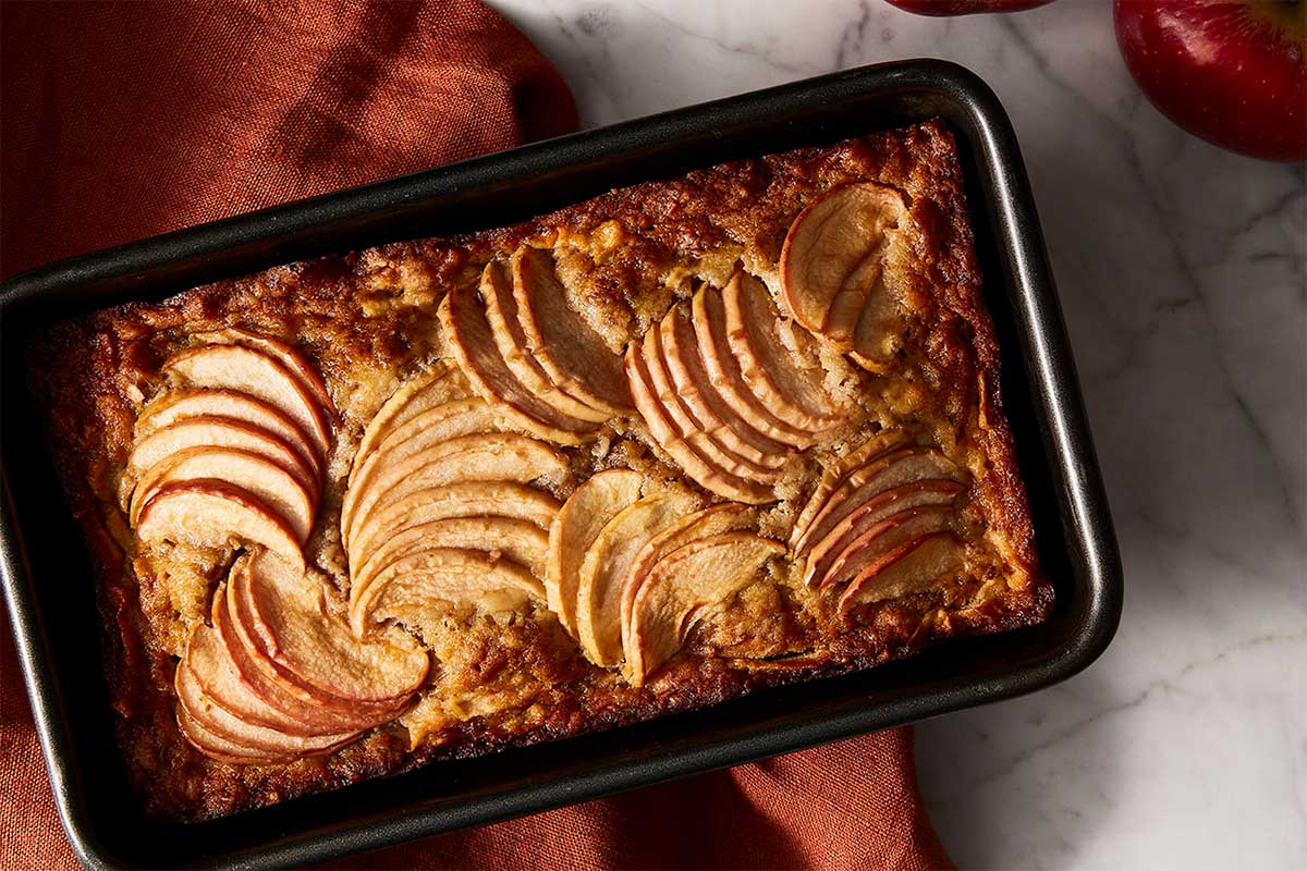 A loaf of cheddar cheese bread in a loaf pan with slices of apples on top