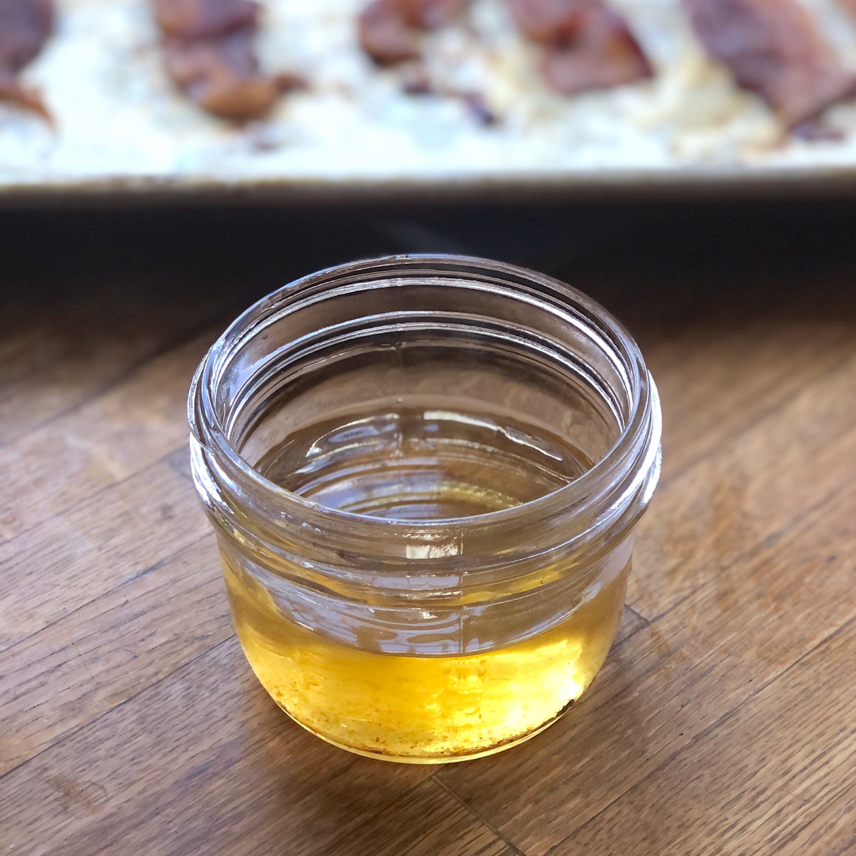 Liquid bacon fat in a small mason jar in front of a baking sheet of baked bacon.