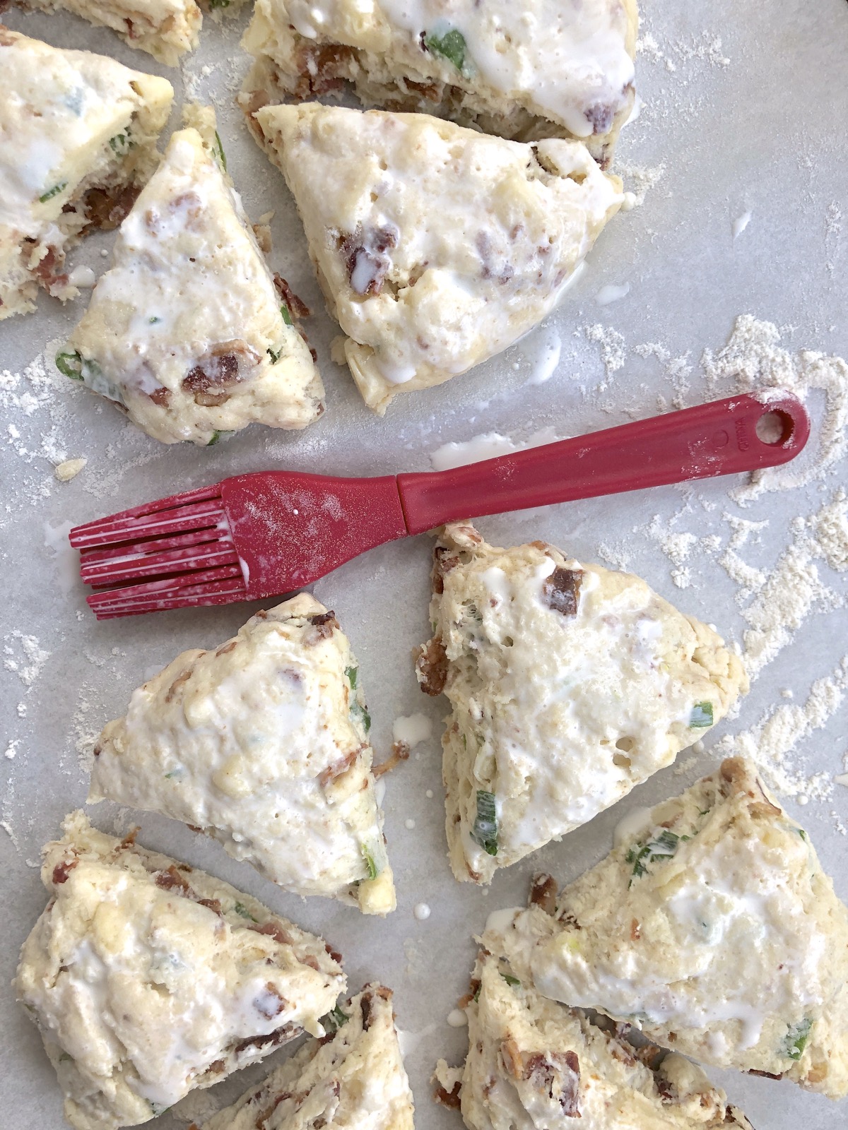 Bacon cheddar chive scones cut into wedges on a baking sheet and brushed with cream using a red silicone pastry brush.