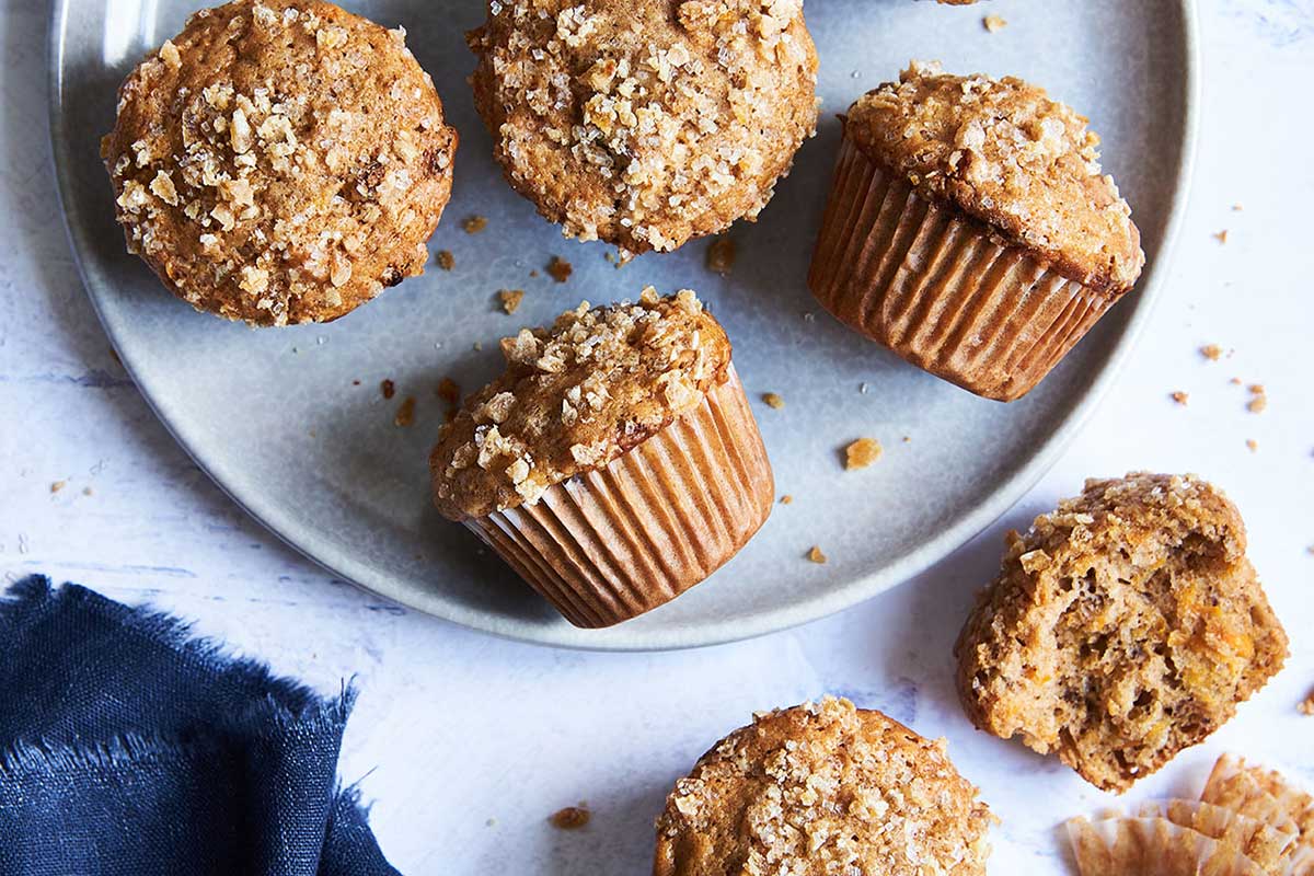 A platter of carrot muffins on a kitchen table with one broken open to show the crumb