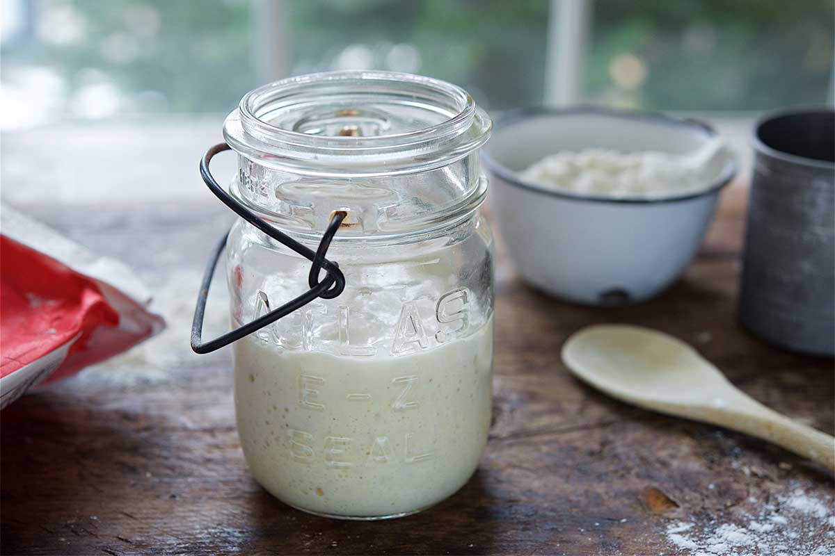 A mason jar of bubbly sourdough starter next to a bag of King Arthur Flour