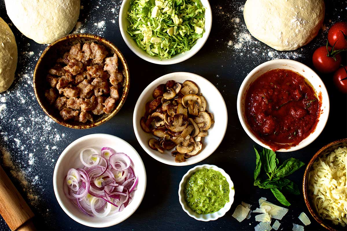 A half dozen small bowls filled with different pizza toppings