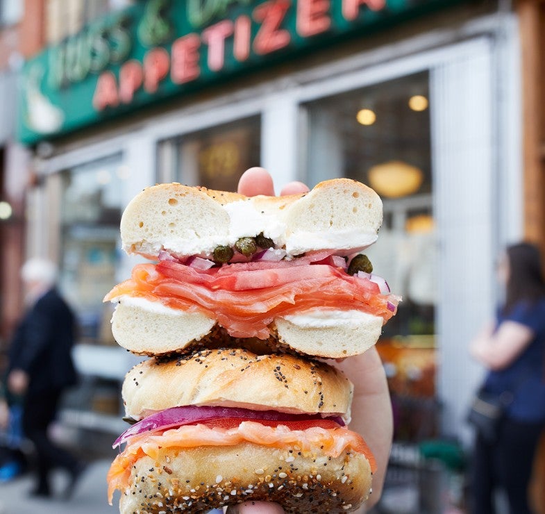 Hand holding sliced lox and bagel sandwich in front of Russ and Daughters storefront