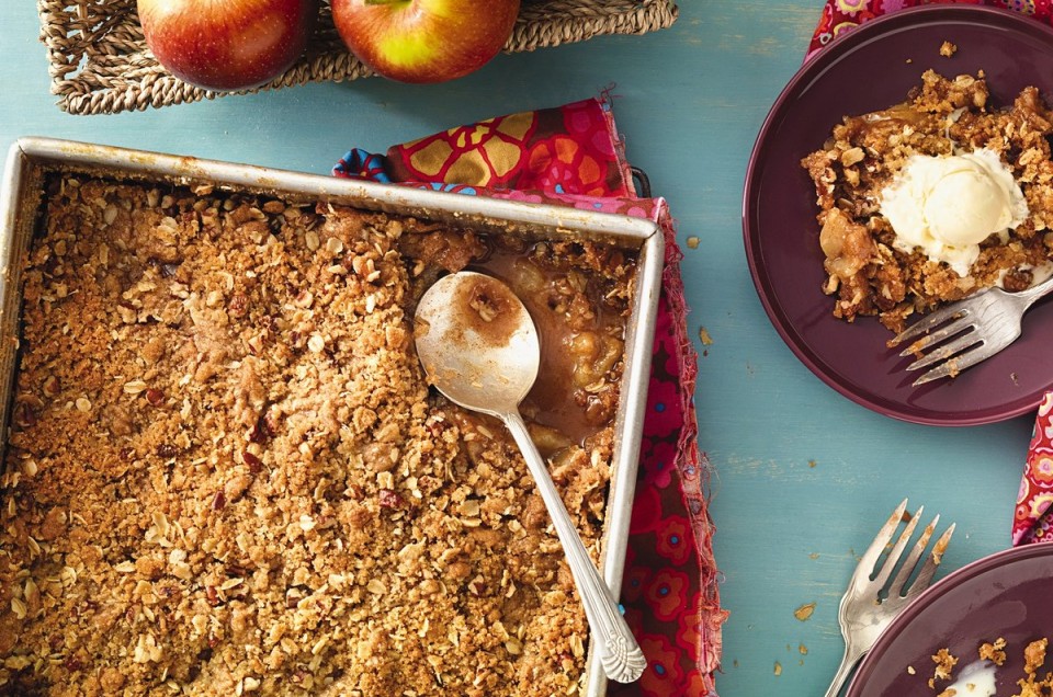 A pan of apple crisp on a table with a few apples and two plates of apple crisp with ice cream