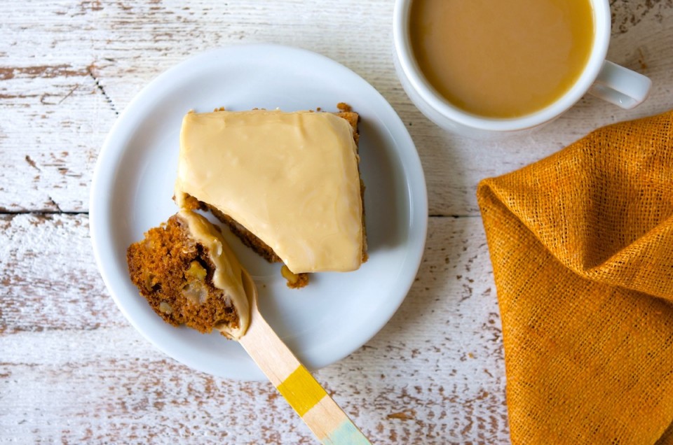 Square of apple cake with brown sugar frosting on a plate with a fork, coffee on the side.