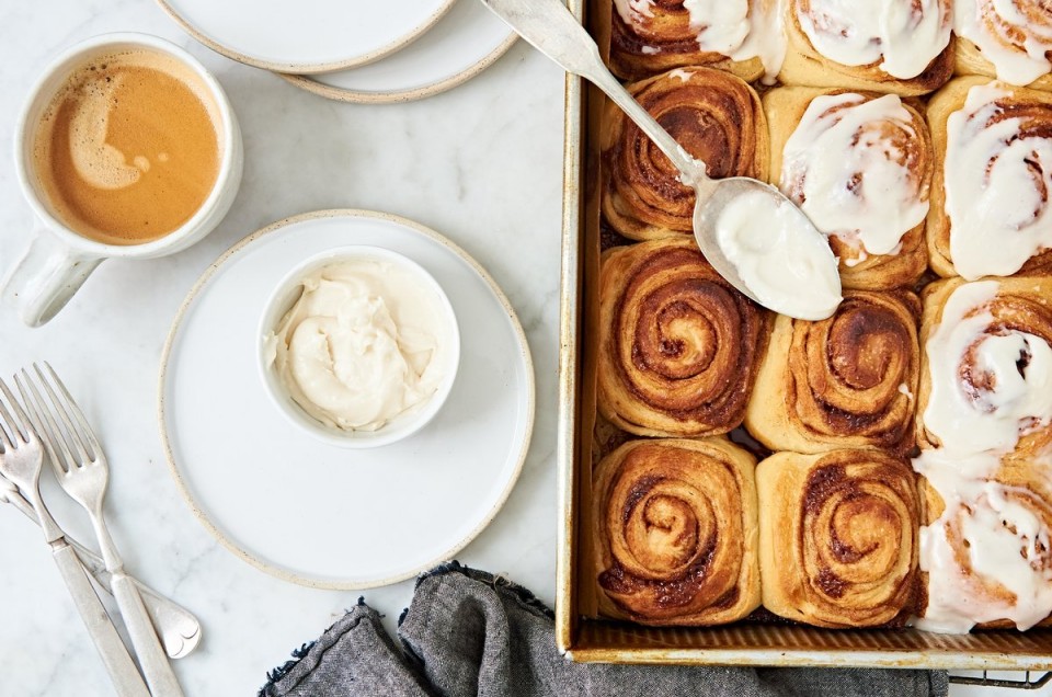 A tray of cinnamon buns with a bowl of frosting next to a cup of coffee