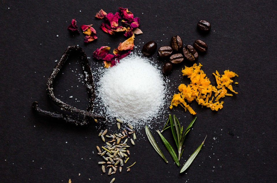 A pile of sugar with ingredients that can be infused into it to add flavor: vanilla bean, rose petals, coffee beans, orange zest, rosemary springs, and lavender flowers