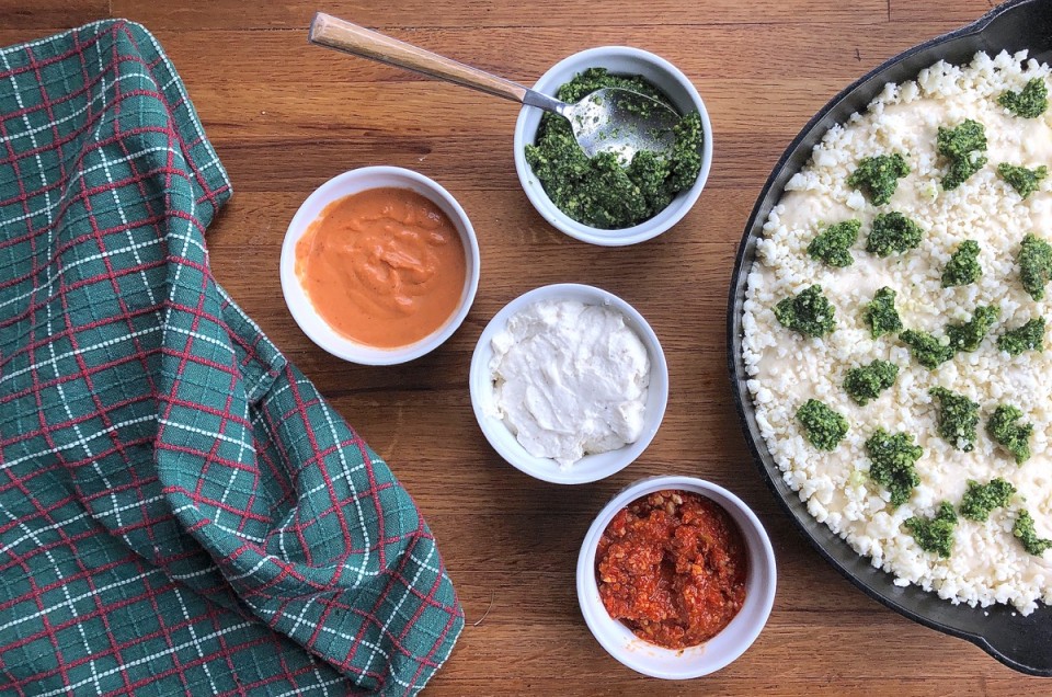 Pizza being topped with sauce, four bowls of sauce: pesto, romesco, white, and pink vodka.