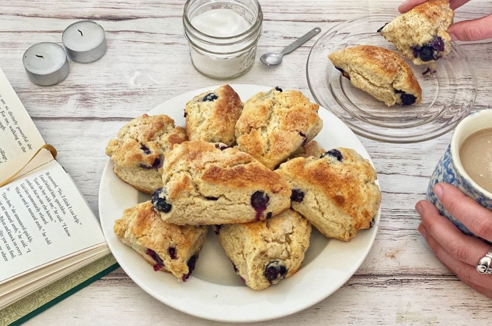 A plate of cream scones next to a book and a person holding a mug of coffee