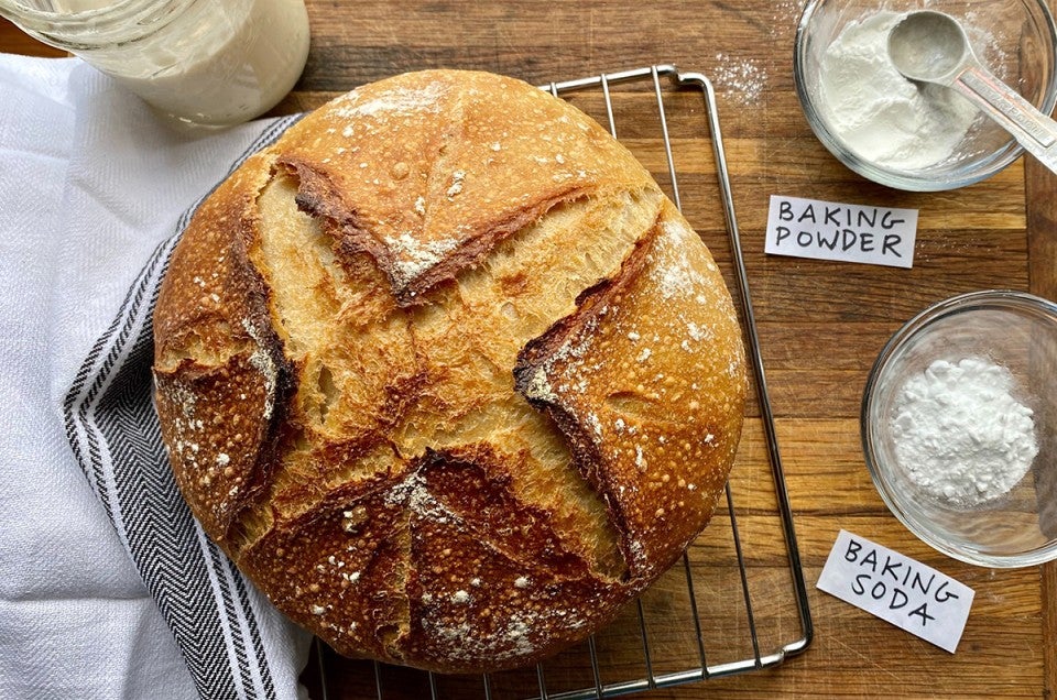 A loaf of sourdough bread on a cooling rack next to a starter and a bowl of baking powder and baking soda