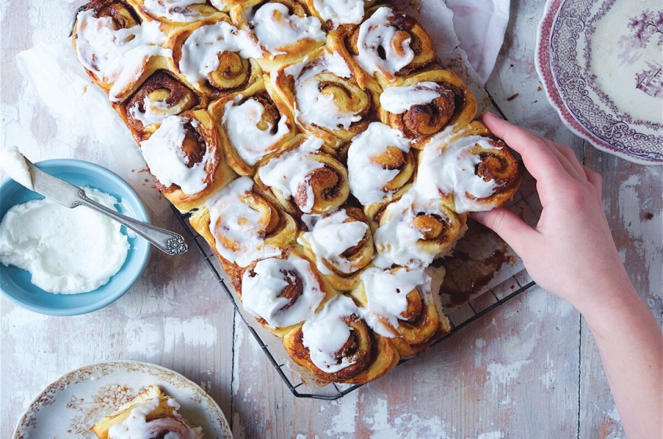 A batch of soft cinnamon rolls on a cooling rack with a hand reaching for a roll