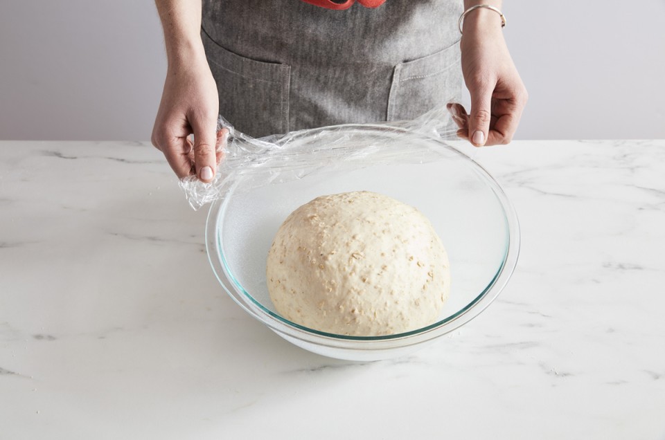 Bread dough in a bowl being covered with plastic wrap
