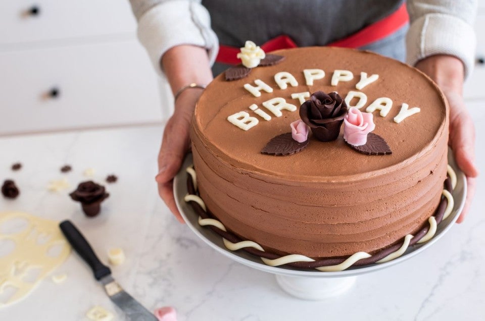 A baker presenting a birthday cake topped with modeling chocolate letters and flowers