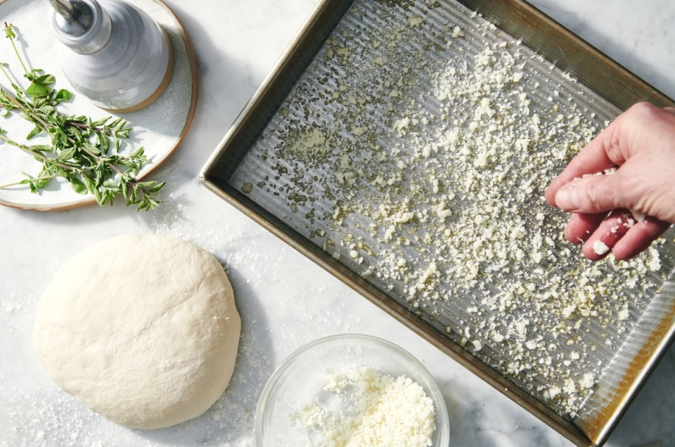 Grated Paremsan being sprinkled into an oiled pizza pan before adding the dough on top.