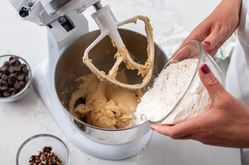 Hands adding flour to a stand mixer to make cookie dough