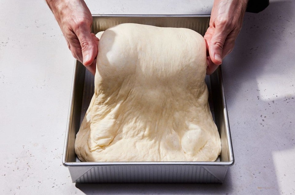 A baker folding wet dough in a square pan with their hands