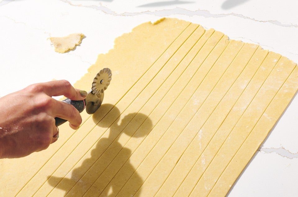 Rolled out gluten-free pasta on a counter showing a hand with a pasta cutter cutting strips. - select to zoom