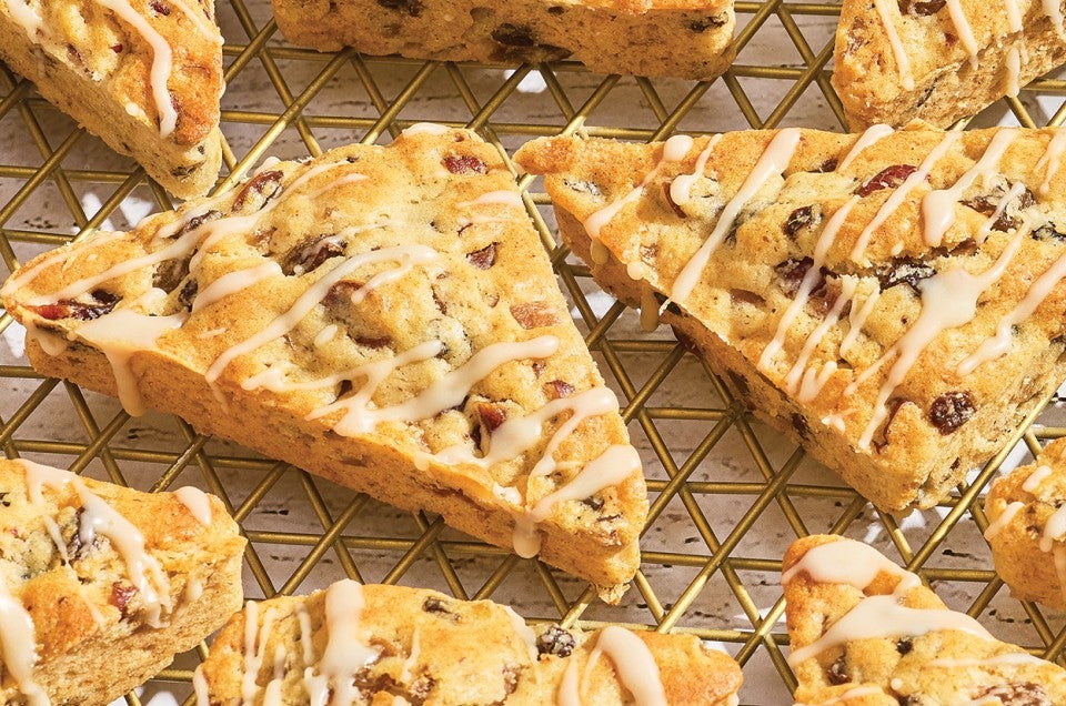 Multiple fruitcake scones photographed on a cooling rack showing golden brown scones with a glaze drizzled atop and dotted with dried fruit. - select to zoom
