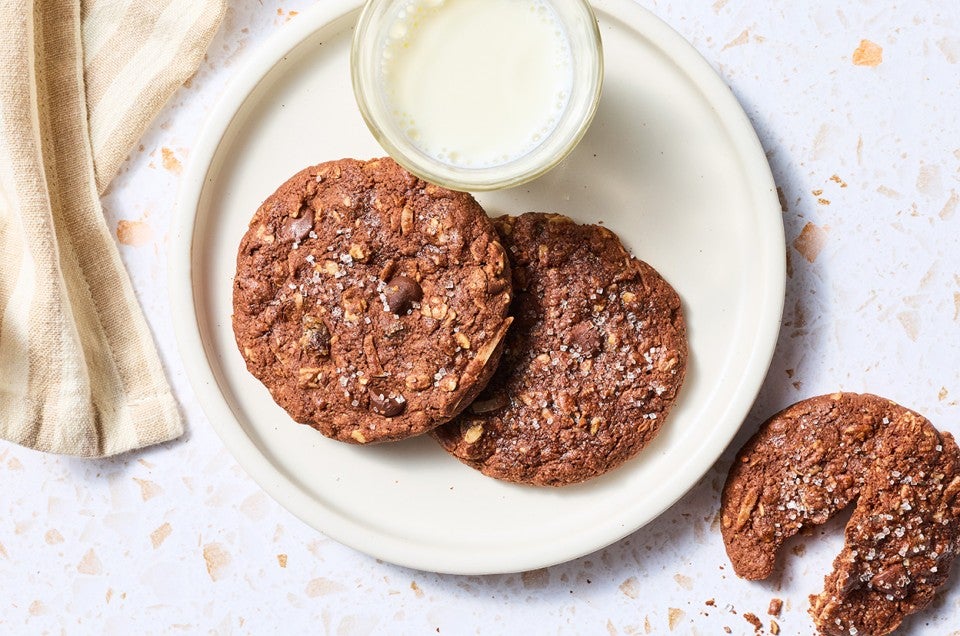 Two Chocolate Lactation Cookies on a plate with a glass of milk and another cookie on the counter next to the plate. - select to zoom