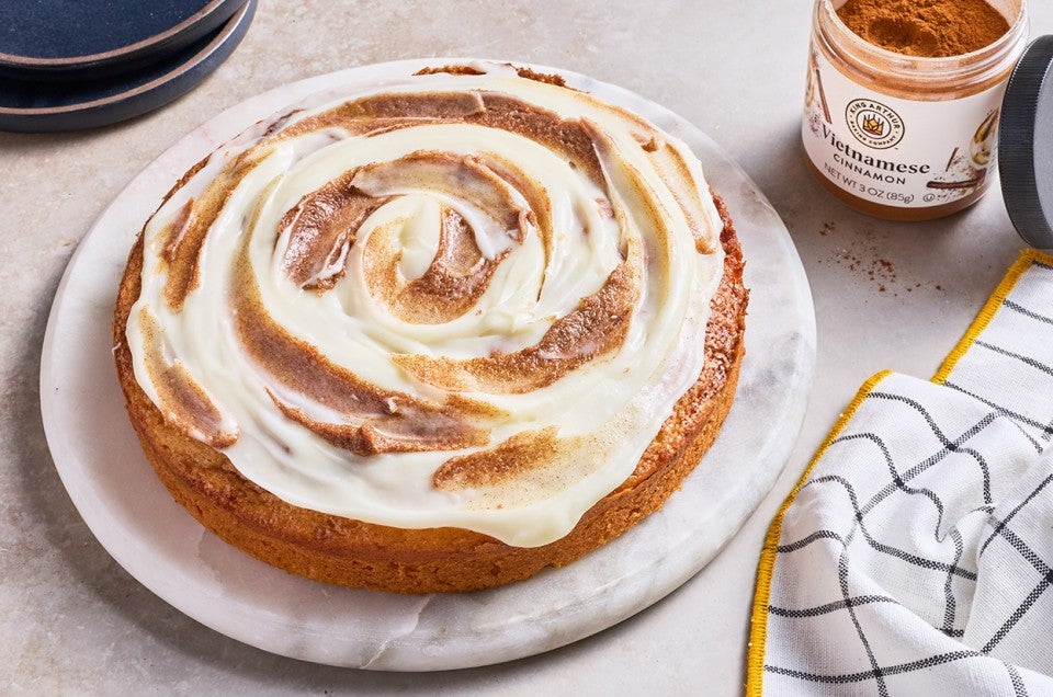 Cinnamon Roll Cake on a marble round cake platter and a jar of Vietnamese cinnamon in the background. - select to zoom