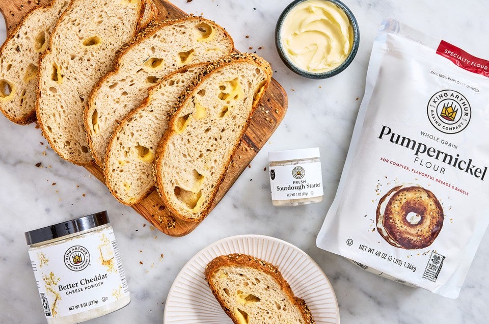 Parmesan Black Pepper Sourdough Bread sliced on a wooden cutting board with butter, a jar of sourdough starter, better cheddar cheese powder, and pumpernickel flour. - select to zoom