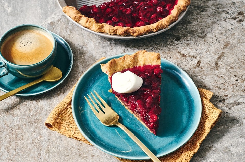 A slice of Cranberry-Fudge Pie topped with a dollop of whipped cream, a cup of coffee sits on a plate off to the side. - select to zoom