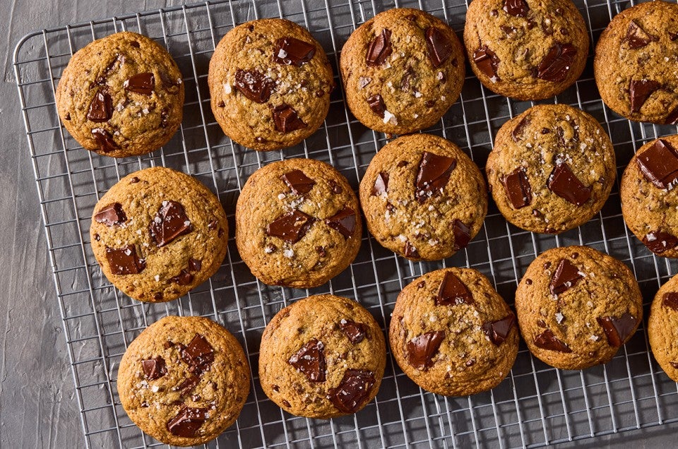 Several beautifully baked High-Altitude Chocolate Chip Cookies shown on a plate and counter with a glass of milk in the background. - select to zoom