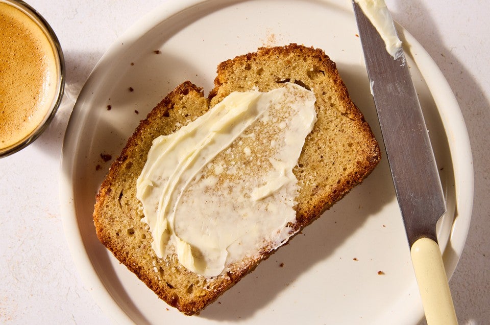 A slice of High-Altitude Banana Bread buttered and laying on a light plate with a butter knife laid on it's side next to the bread. - select to zoom