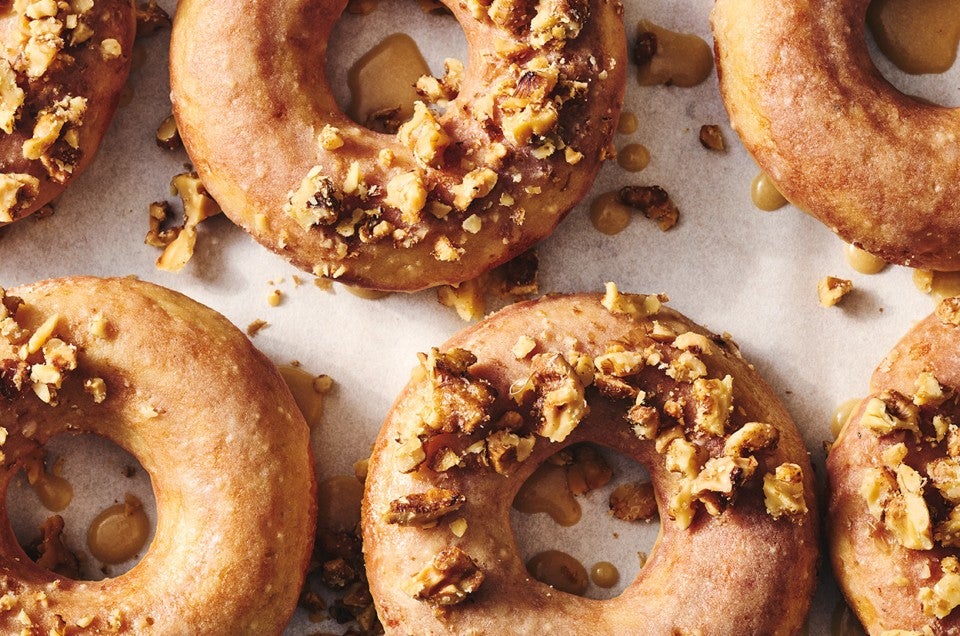 Multiple Maple Donuts on a sheet tray with half coated in broke nut meat and the other half glazed. - select to zoom