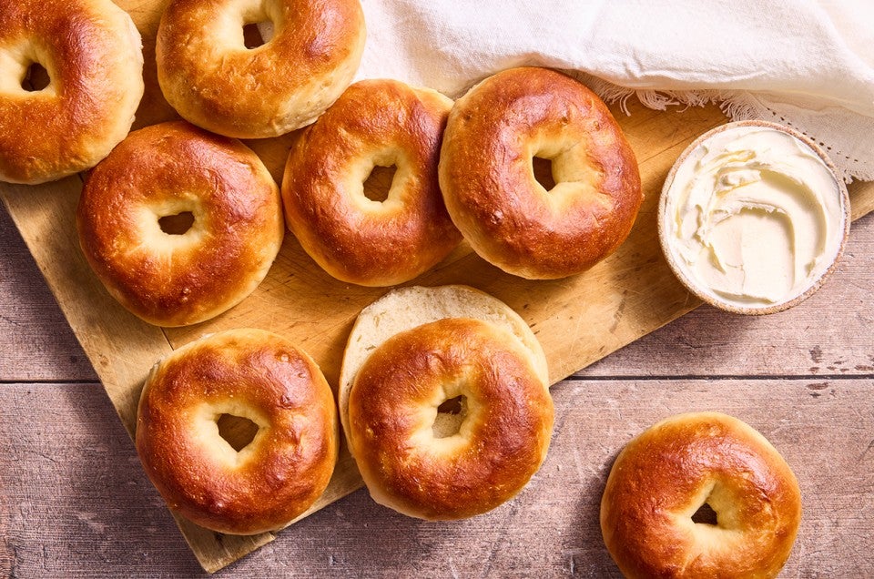 Several bagels laid out on a cutting board with a tub of cream cheese in the upper right corner of the frame. - select to zoom