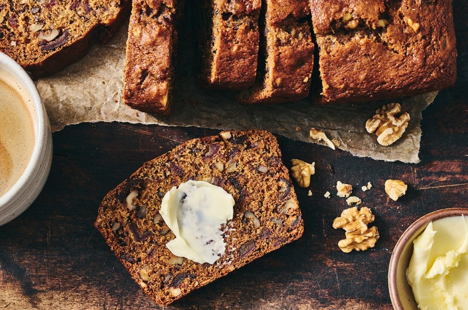 A slice of buttered Old-Fashioned Date Nut Bread on a plate atop a dark counter and with a latte and sliced loaf in the corner of the photo. - select to zoom