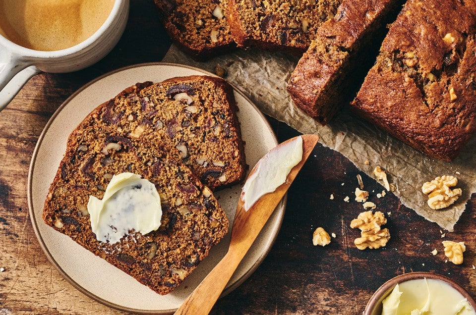 A slice of buttered Old-Fashioned Date Nut Bread on a plate atop a dark counter and with a latte and sliced loaf in the corner of the photo. - select to zoom