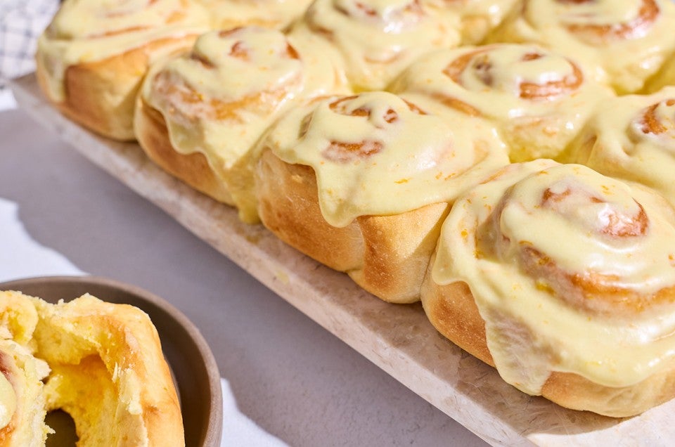 Fluffy Frosted Orange Rolls on a sheet tray showing the side of the rolls with a golden brown and a glossy yellow orange glaze coating them. - select to zoom