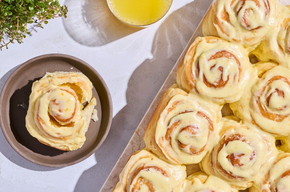 Fluffy Frosted Orange Rolls seen from above on a sheet tray with a single roll on a dark plate. - select to zoom