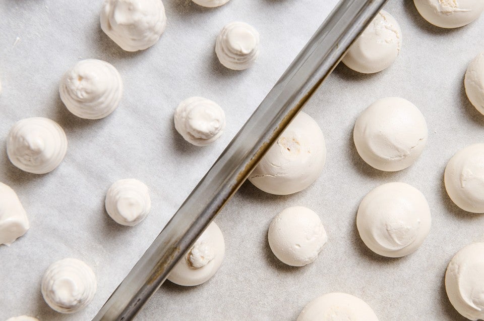 Swiss Meringue piped into round balls on sheet trays atop parchment paper ready for the oven. - select to zoom