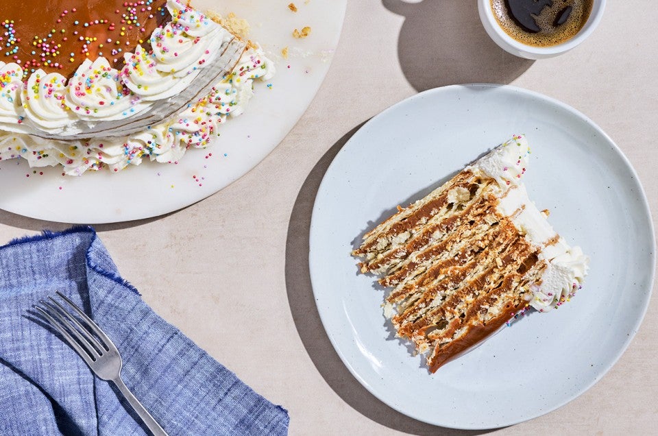 Overhead photograph of Torta Chilena sliced and laying on its side on a white plate. Layers of dulce de leche, flaky pastry, and creme fraiche frosting can clearly be seen. - select to zoom
