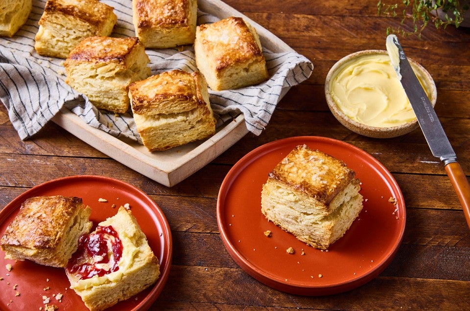 A tray of tall flaky biscuits, a plate with a single rectangular biscuit, a bowl of butter, and another red plate with a sliced biscuit that has jelly and butter on it. - select to zoom