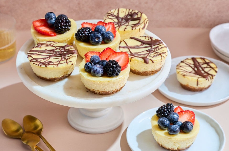 Mini Cheesecakes arranged on a pedestal and plates with fresh berries and chocolate drizzle topping them. - select to zoom