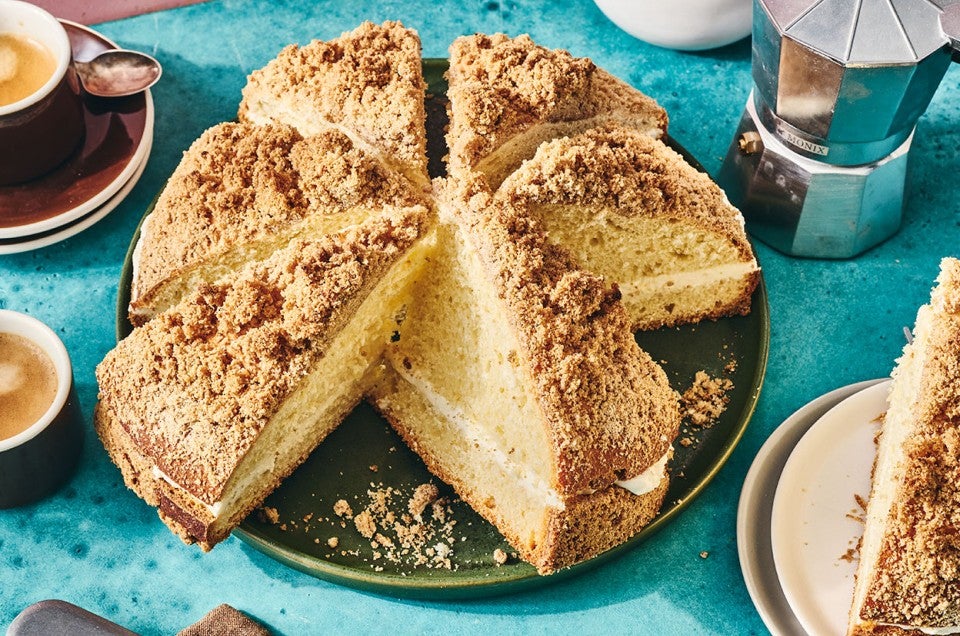 A round, Cream-Filled Coffee Cake, sliced on a dark plate with other breakfast elements spread around a table. - select to zoom
