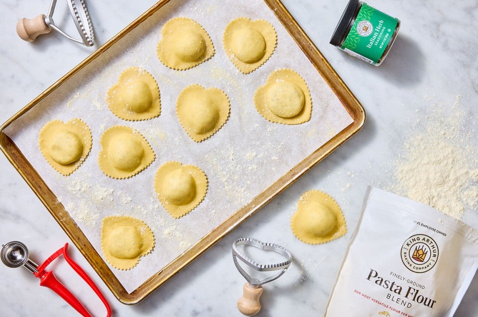 Homemade Cheese Ravioli shown cut into heart shapes and organized on a sheet tray covered in parchment with ingredients and tools strewn about a light marble counter. - select to zoom