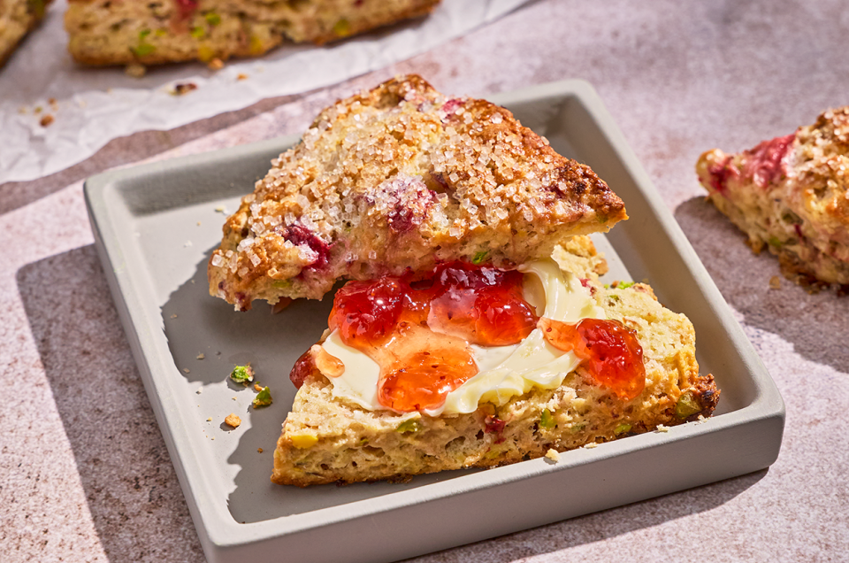 A Roasted Strawberry and Pistachio Scone sliced in half with cream and dollops of strawberry preserve. - select to zoom