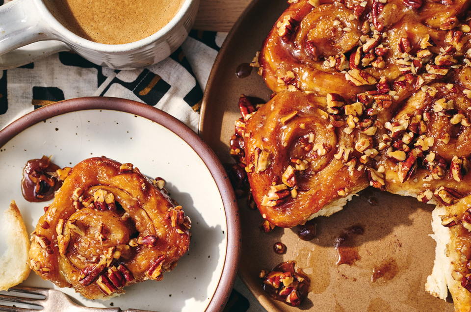 A single sticky bun on a plate adjacent to freshly baked buns. - select to zoom