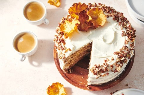 An overhead photo of a Hummingbird Cake with pineapple cream cheese frosting. A slice has been taken out of the cake and there are toasted nuts and a slice of dried pineapple on top of the cake.  - select to zoom