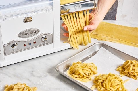 Cutting pasta into noodles with an electric pasta machine, next to a sheet pan of fresh pasta noodles