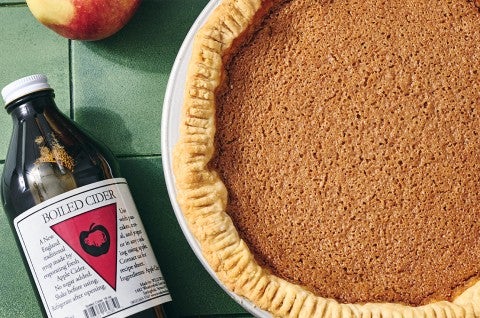 An apple pie seen from above with a smooth texture and a bottle of boiled cider laying on the table next to it. - select to zoom