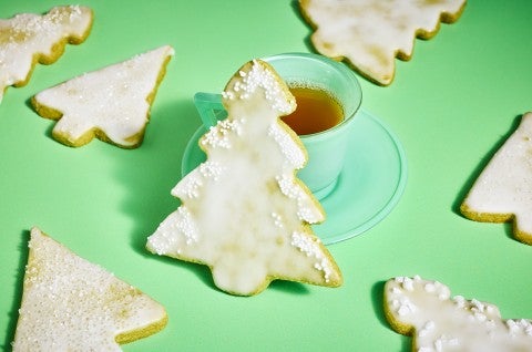 A matcha Christmas tree cookie leaning against a cup of tea with other cookies spread out on a green table cloth. - select to zoom